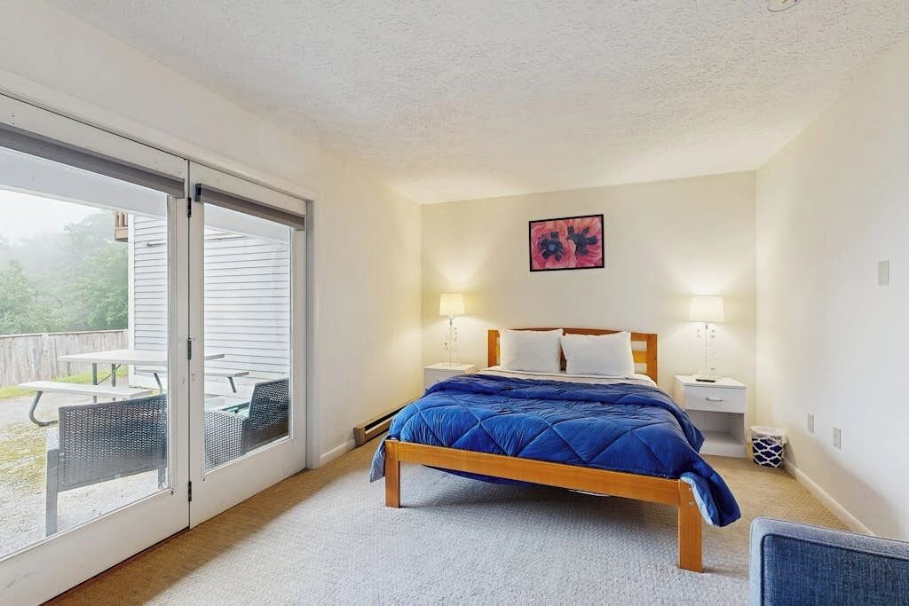 Un dormitorio con una cama, dos mesitas de noche, una mesita auxiliar, una puerta corrediza de vidrio a un patio y un cuadro colgado en la pared.