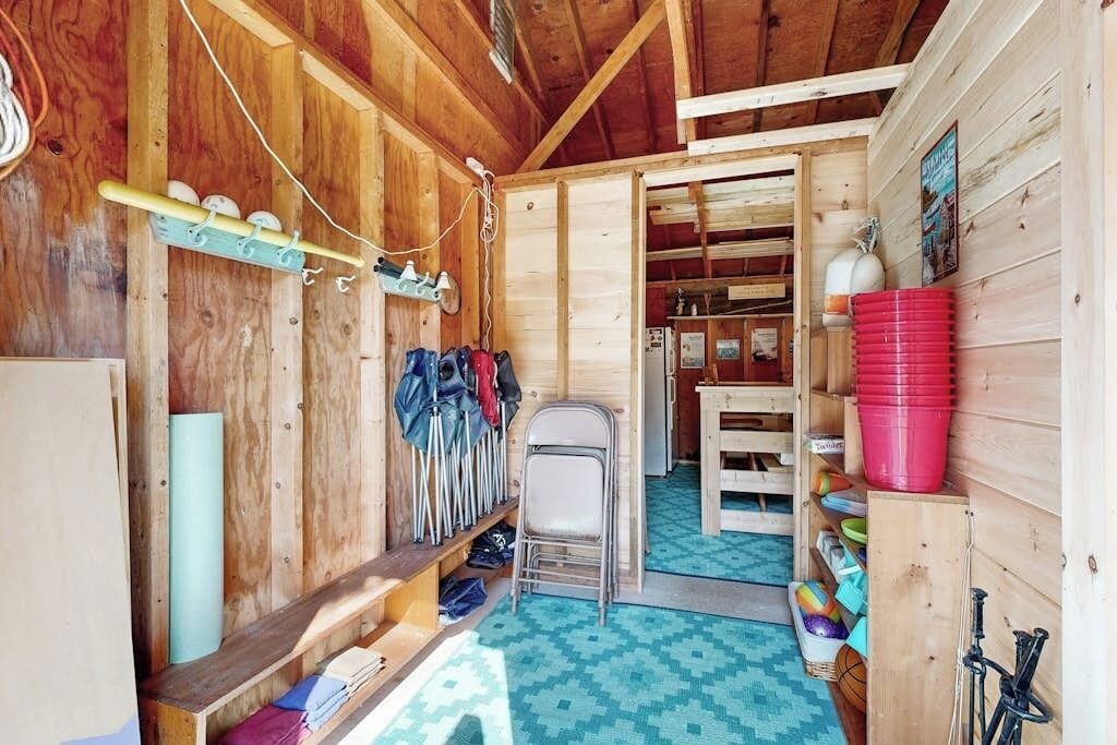 Interior d'una cabana de fusta amb un banc de fusta, una cadira plegable, prestatges amb diversos objectes i una porta que condueix a una altra habitació.