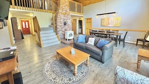 A spacious living area with a high ceiling, a fireplace, a staircase, and a dining area. - Riverfront Home w/ Deck, Amazing Views & Firepit - Near the Best of Maine (Stockton Springs)