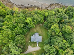 An aerial view of a property with a house surrounded by dense forest, a clear lawn, and a beach in the background. - Riverfront Home w/ Deck, Amazing Views & Firepit - Near the Best of Maine (Stockton Springs)