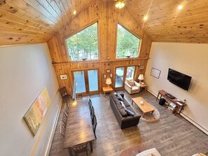 A spacious room with a wooden ceiling, large windows, a sofa, a dining table, and a television. - Riverfront Home w/ Deck, Amazing Views & Firepit - Near the Best of Maine (Stockton Springs)