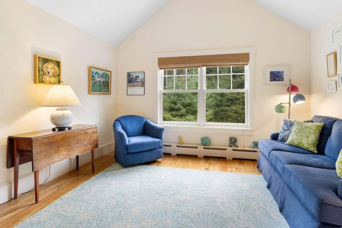 A living room with a sofa, armchair, and a table with a lamp. There is a window with a view of greenery outside.
