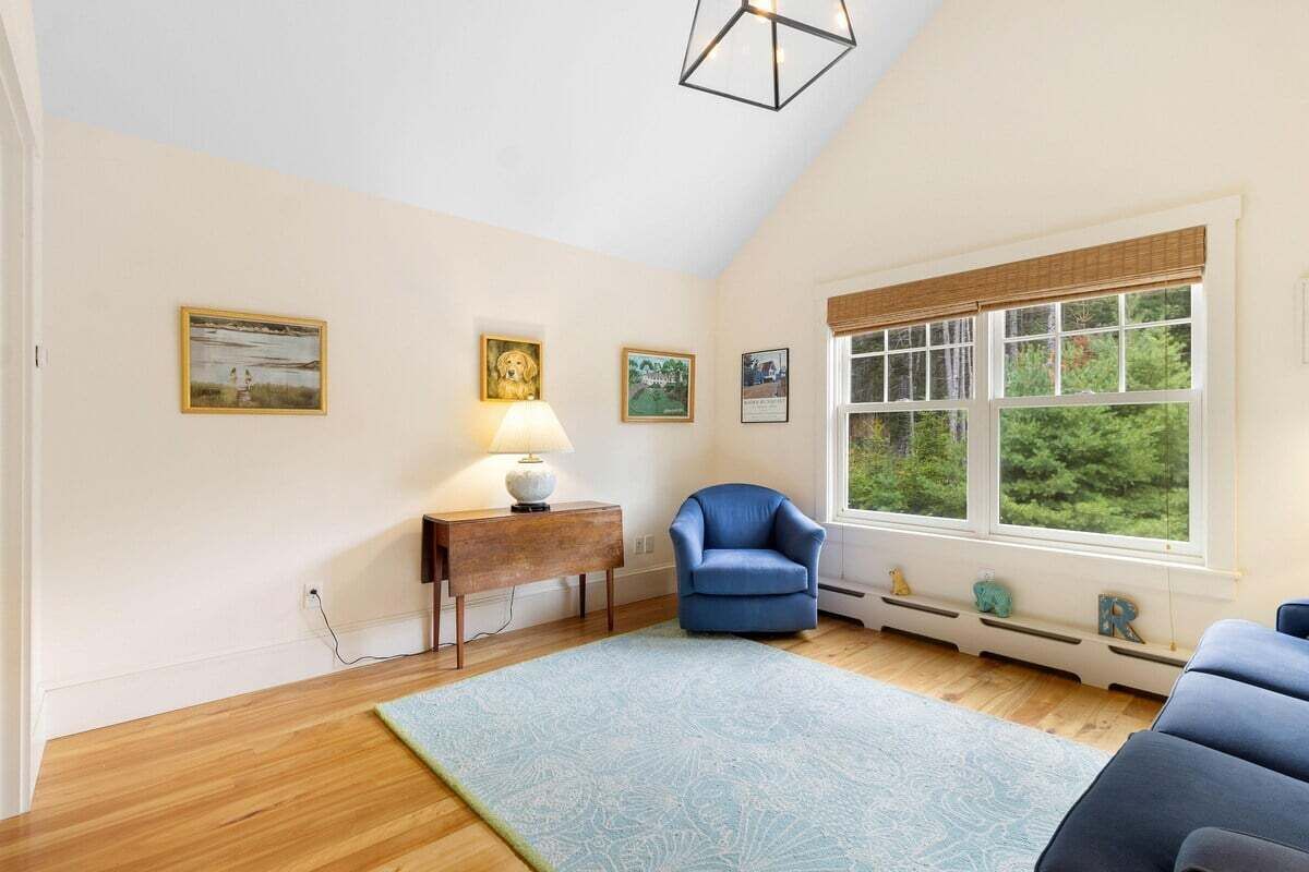 A room with a wooden floor, a blue armchair, a small table with a lamp, and a large window with a view of greenery.