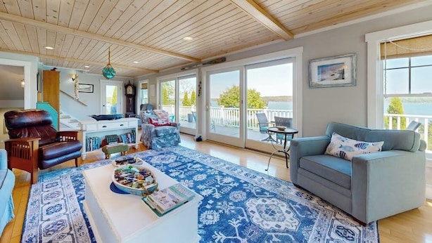 Un salon spacieux doté d’un plafond en bois, d’un tapis à motifs bleus, d’un canapé gris et offrant une vue sur l’extérieur grâce à de grandes fenêtres.