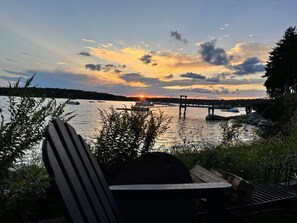 Blick auf einen See mit einem Steg, einem Stuhl und einem kleinen Gebäude, vermutlich eine Hütte oder ein Bootsheim.