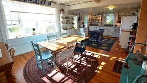 A dining area with a wooden table and chairs, a kitchen in the background, and a window with blinds. - Berry Cottage, Private Dock, Overlooking a Sandy b (South Thomaston)