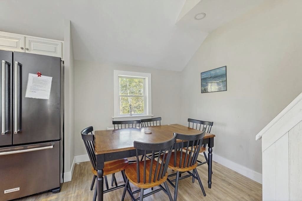 A dining area with a wooden table and chairs, a stainless steel refrigerator, a window with a view, and a painting on the wall.
