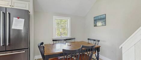 A dining area with a wooden table and chairs, a stainless steel refrigerator, a window with a view, and a painting on the wall.