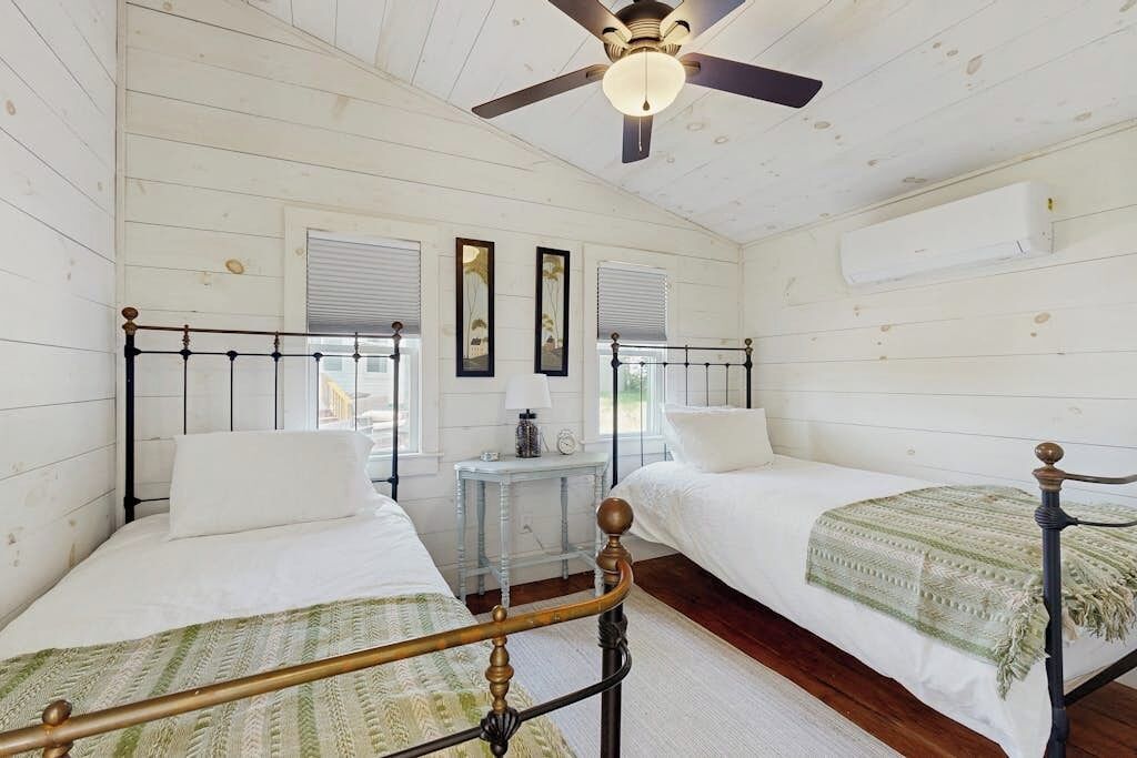A room with two beds, a ceiling fan, a small table, and a window with blinds.