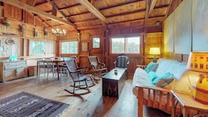 A rustic cabin interior with wooden walls, a dining area, a sofa, rocking chairs, and a lamp. - Waterfront Cabin With Ocean View & Private Dock (East Boothbay)