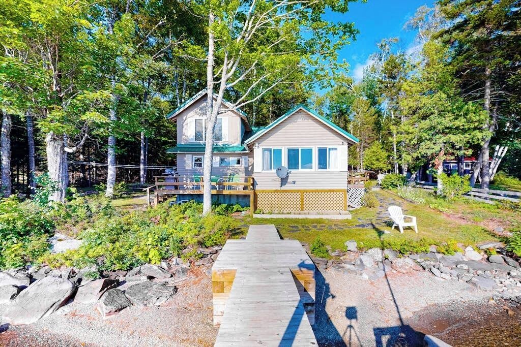 A cabin with a deck, surrounded by trees and a wooden walkway leading to it.