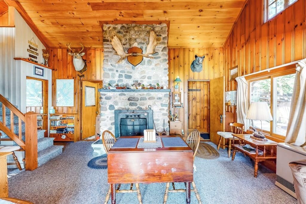 Interior de una cabaña de madera con chimenea de piedra, una mesa de comedor y una cabeza de oso montada en la pared.