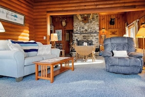 Ein Holzblockhaus-Interieur mit einer Couch, einem Sessel, einem Holztisch, einem Kamin aus Stein und aufgehängten Tierköpfen.