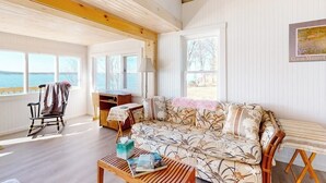 Huone, jossa on puinen katto, kukkapainokuosilla verhoiltu sohva, keinutuoli, lamppuvarustettu pöytä ja näkymä ulos.