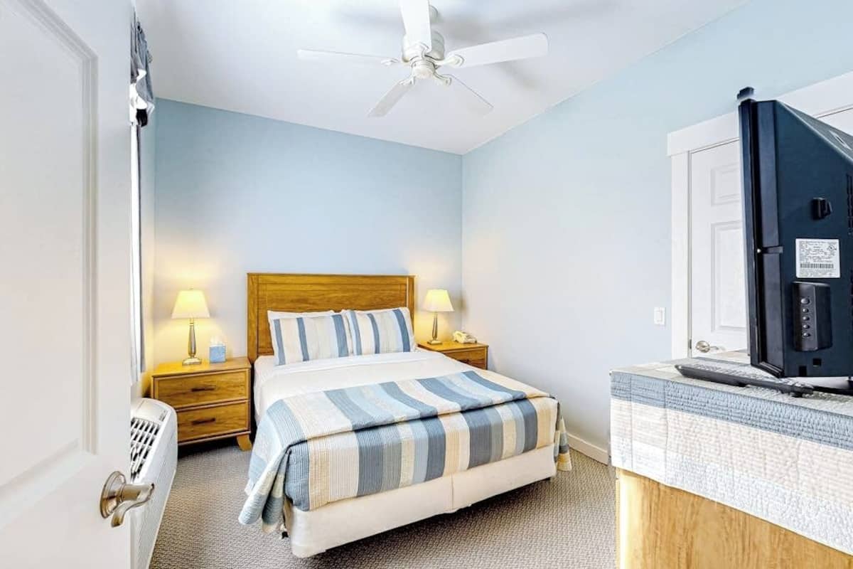 Habitación de hotel con cama, lámparas de noche, escritorio con computadora, ventilador de techo y una puerta.