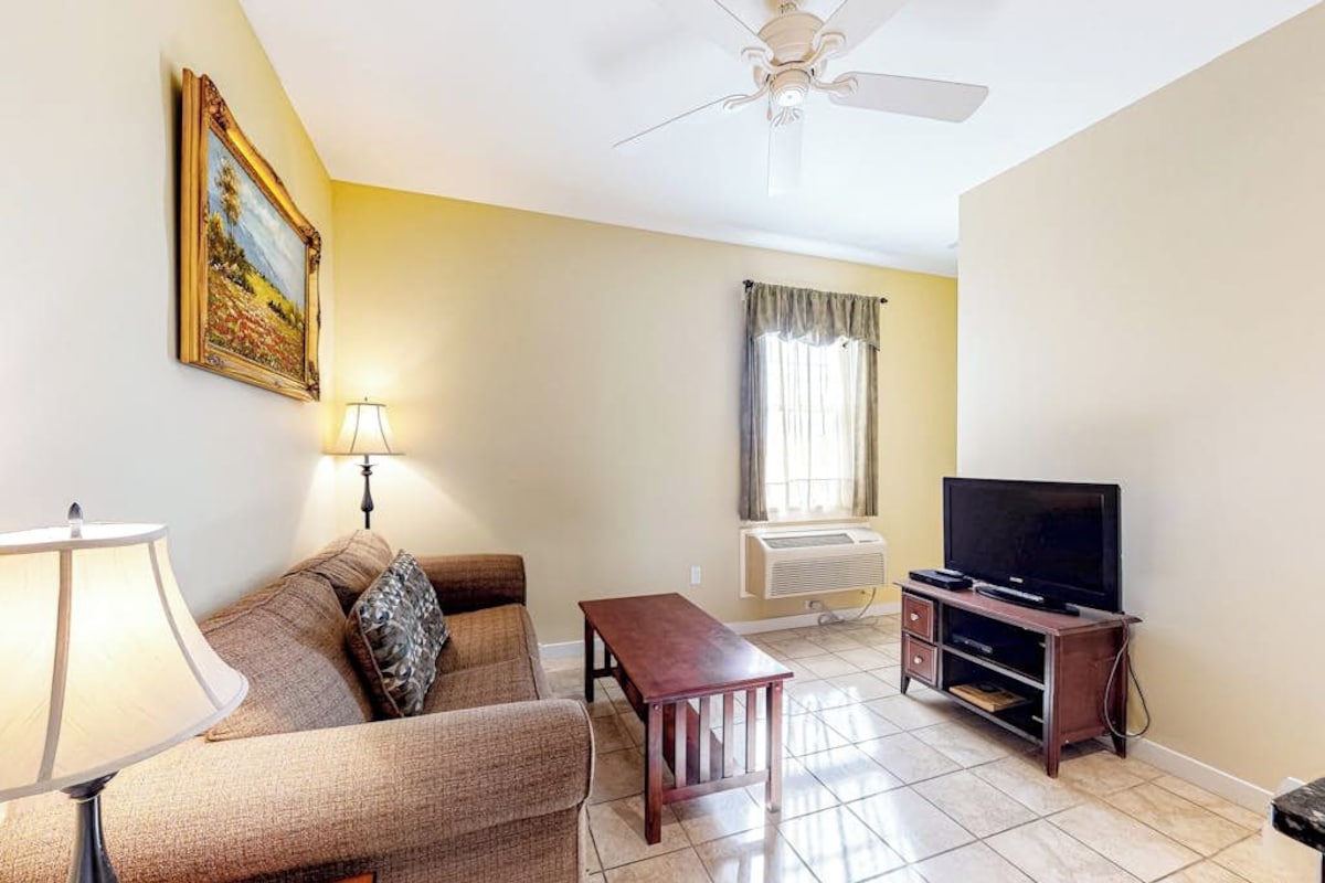 A living room with a sofa, a table, a TV, a ceiling fan, and an air conditioning unit.