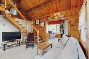 Interior de una cabaña de troncos con televisor, sofá, escalera de madera y una decoración con cabeza de venado.