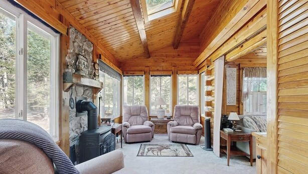Een gezellig interieur van een blokhut met een open haard, twee fauteuils, een bank en een groot raam met uitzicht op het bos.