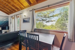 A room with a wooden ceiling, a dining table with chairs, a sofa, a window with a view of trees, and a painting on the wall. - Secluded Sunset Point Mountain top Cabin, Wifi, Dish TV, Firepit (Bar Harbor)