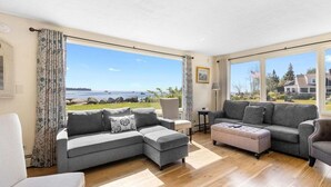 A spacious living room with a large window offering a view of the sea and a house. There are two sofas and a small table. - Owls Nest on Crescent Beach (Owls Head)