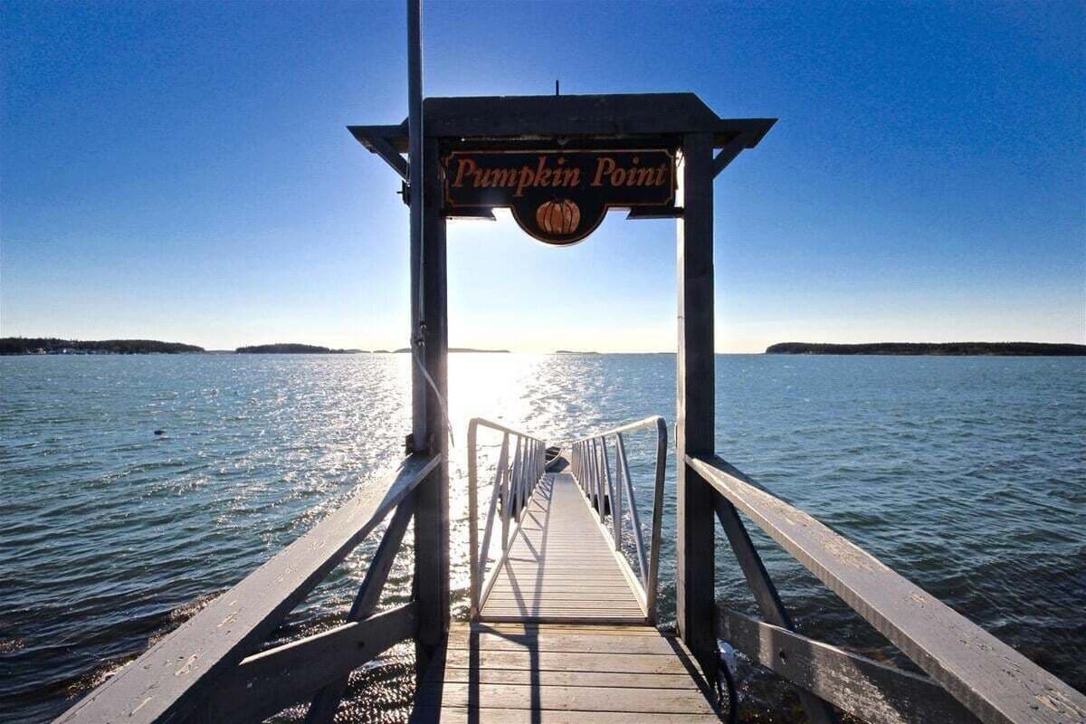 Un muelle de madera que conduce a un cartel que dice 'Pumpkin Point', con vista a un agua tranquila y terreno lejano.
