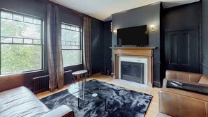 A modern living room with a fireplace, a flat-screen TV mounted above it, and a large window with curtains. - Beautiful Home With Ping-pong, Fast Wifi & W/D - Walk to Dining (Wiscasset)