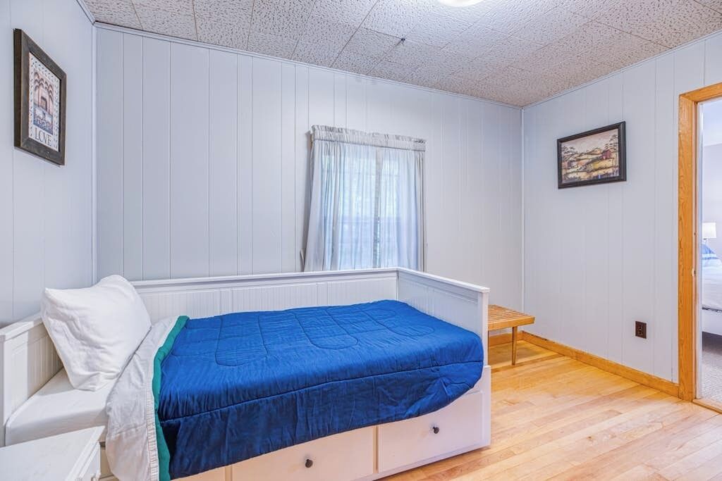 Ein Schlafzimmer mit einem Bett, einem kleinen Tisch, einem Fenster mit Vorhängen und einem Gemälde an der Wand.