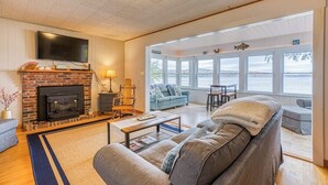 A living room with a fireplace, a television, and a view of the water. - Lakefront Cottage w/ Beautiful Views of Moosehead Lake & Big Moose Mountain (Greenville)