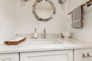 A bathroom with a white marble sink, a round mirror, a towel rack, and a wicker basket. - Elegant Home With Veranda & Water View, Walk to Downtown Boothbay Harbor (Boothbay Harbor)