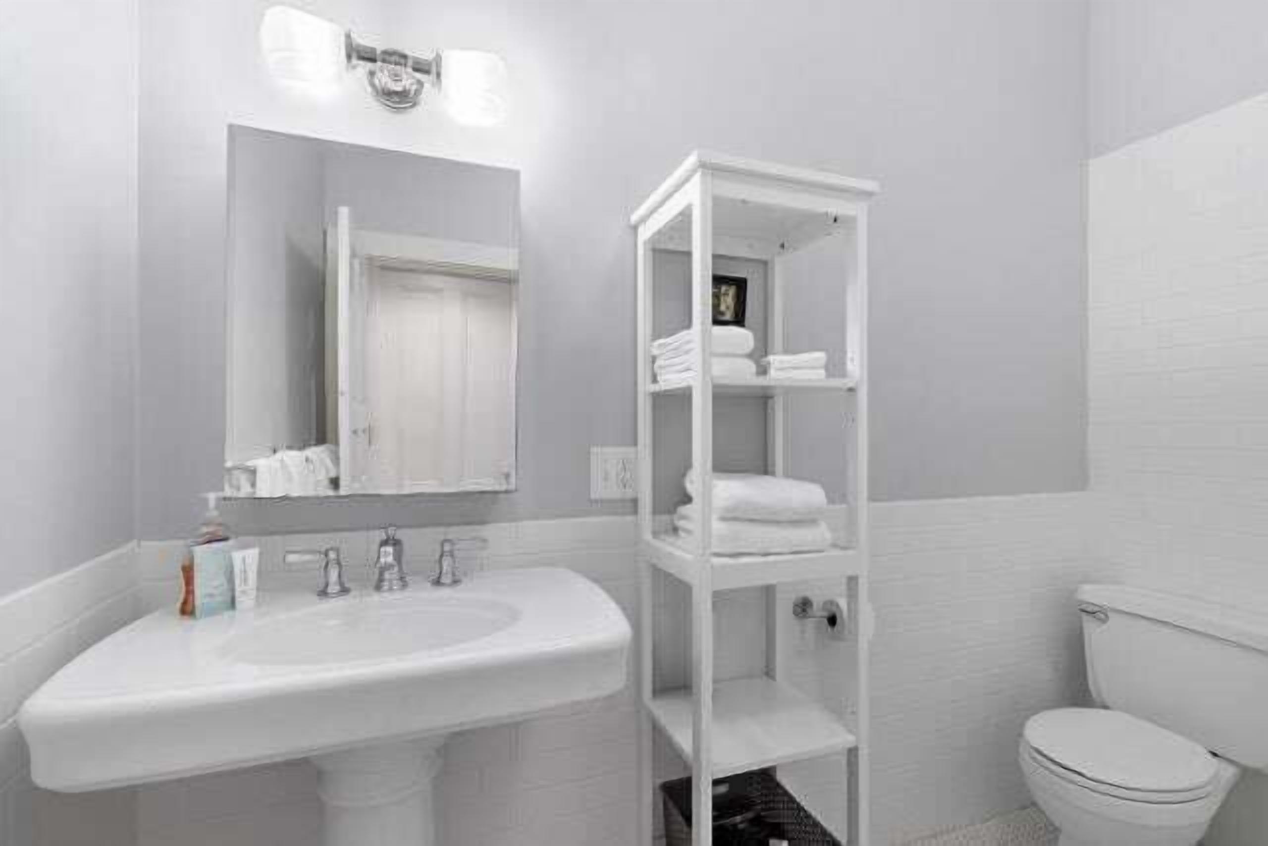 A bathroom with a white sink, mirror, and toilet. There is a white shelving unit with folded towels.
