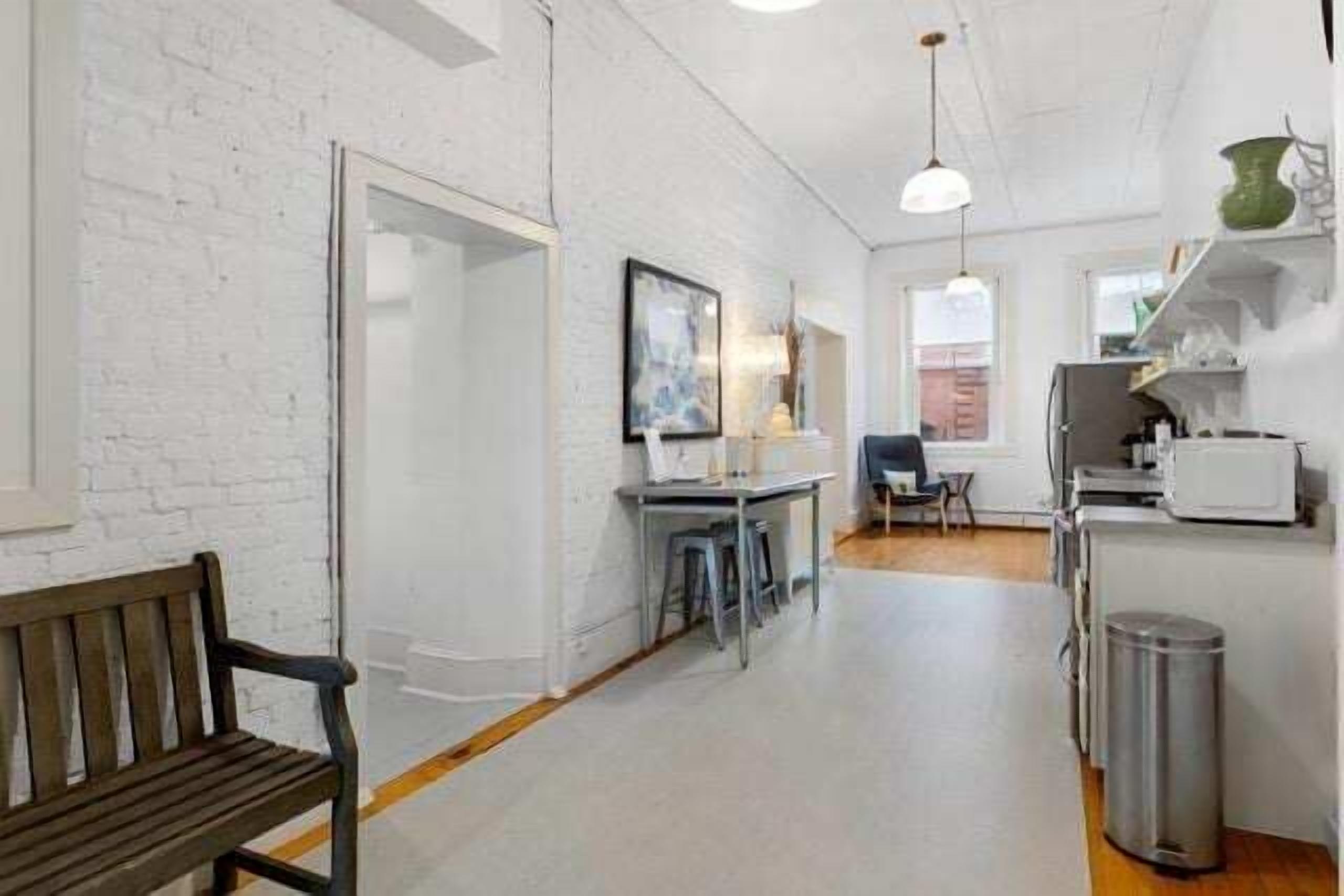 A narrow hallway with a bench, a table with chairs, a refrigerator, and a trash can.