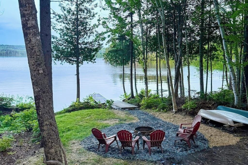 Un espace de détente aménagé au bord du lac, avec des chaises et un foyer, entouré d’arbres et d’un bateau.