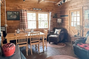 Innenansicht einer Holzhütte mit Esstisch, Stühlen, einer Couch, einer Lampe und einem Schild mit der Aufschrift 'EAT - SLEEP - FISH'.