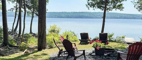 A lakeside seating area with Adirondack chairs, a fire pit, and a view of the lake.