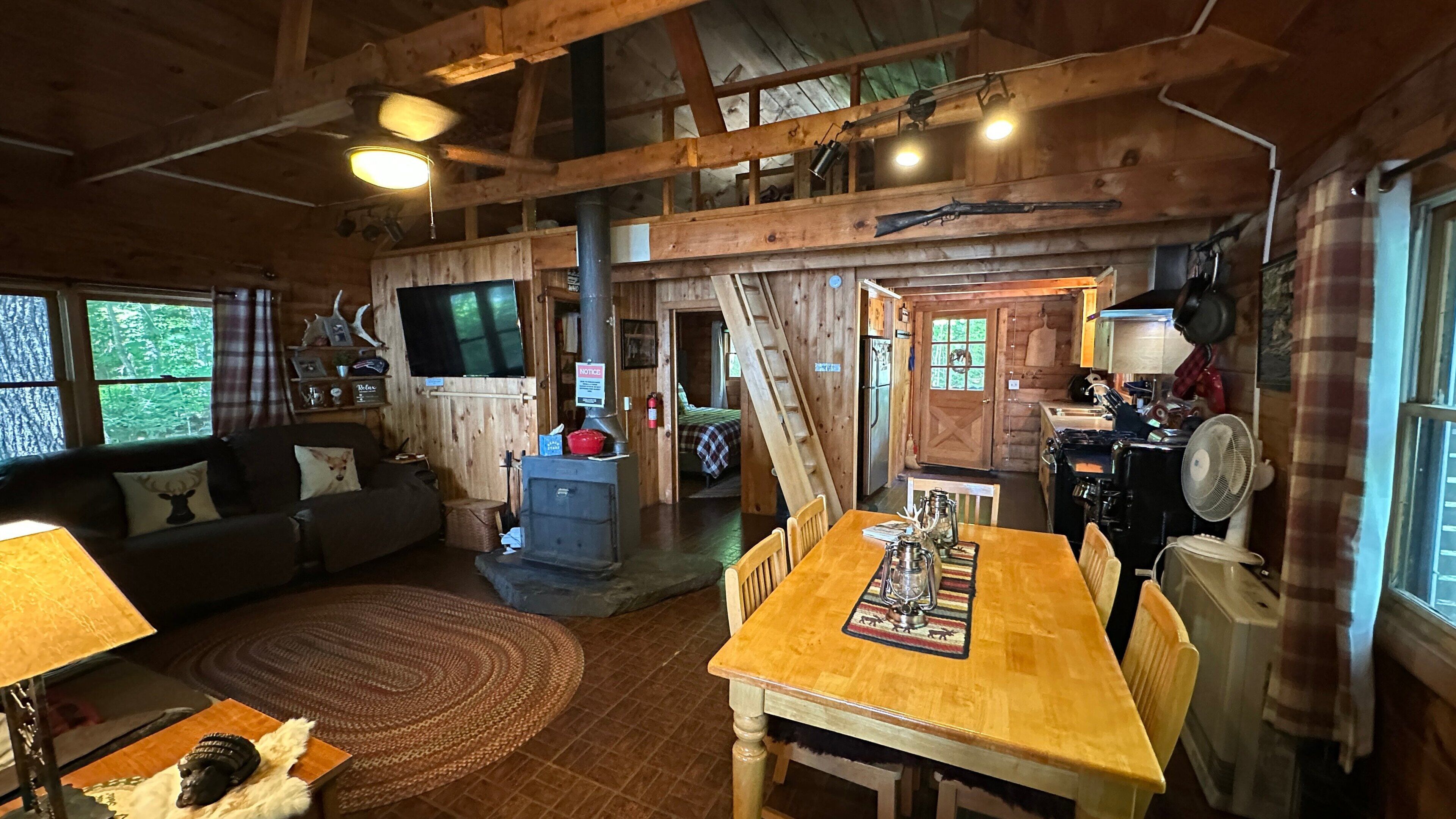 Interior rústico de una cabaña con una mesa de comedor de madera, un sofá, una chimenea y una zona de cocina.