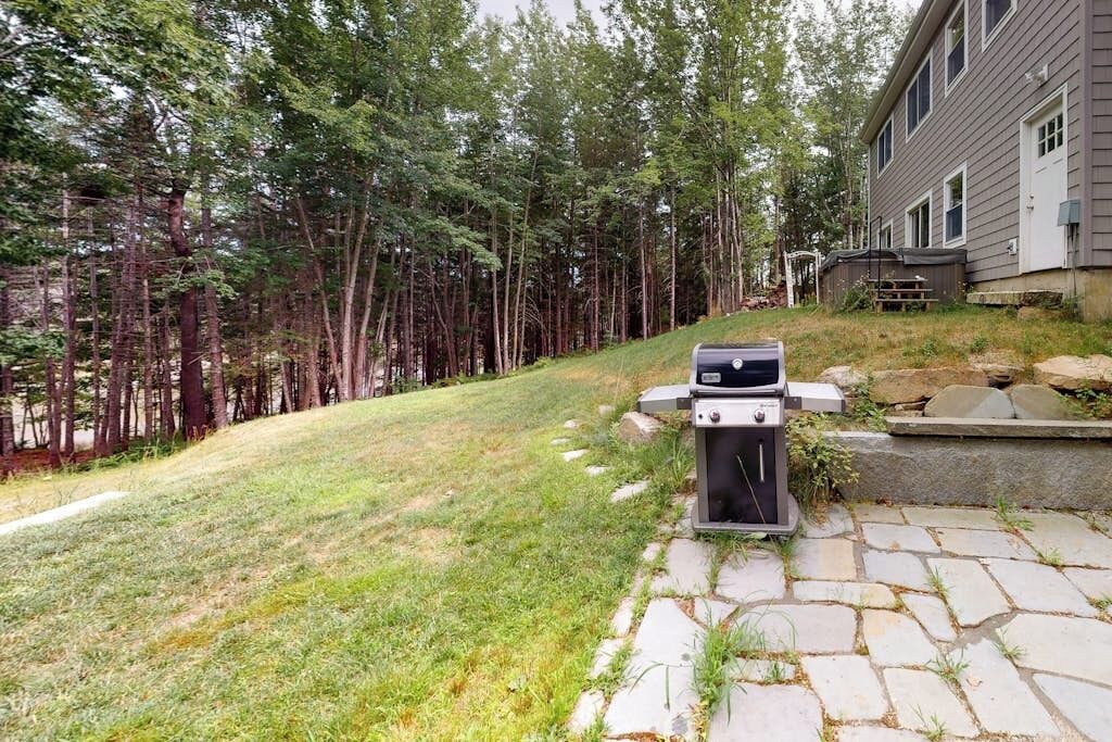Een achtertuin met een pad van stenen dat naar een barbecue leidt, omgeven door een bebost gebied en een huis.