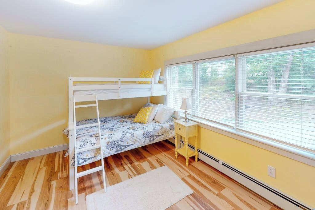 Une chambre avec un lit superposé, une table de chevet avec une lampe, une fenêtre avec des stores et un radiateur.