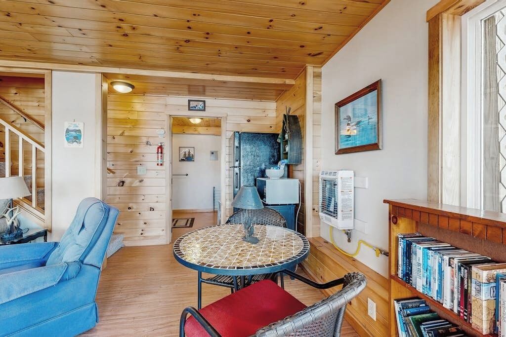 Una habitación acogedora con techo de madera, una mesa redonda con una silla, un sofá azul, una estantería y una puerta que da a otra habitación.
