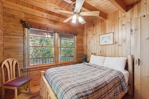 Ein Schlafzimmer einer Holzhütte mit einem Bett, einem Stuhl, einem Deckenventilator und einem Fenster mit Blick auf Bäume.