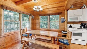 A wooden cabin interior with a dining table, benches, a chandelier, a kitchen with a stove, and large windows. - Dog-friendly Lakefront Home With Deck, Grill, Wifi, Shared Dock & Water Access (Greenville)