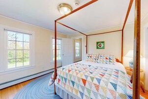 A four-poster bed with a colorful quilt, a round rug, and a view of the outdoors through large windows.