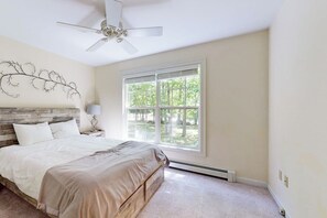 A bedroom with a bed, a nightstand, a ceiling fan, a window with a view of trees, and a wall with a decorative design. - Peaceful Home With Forest Views & Great Yard - Minutes to Town (Wiscasset)