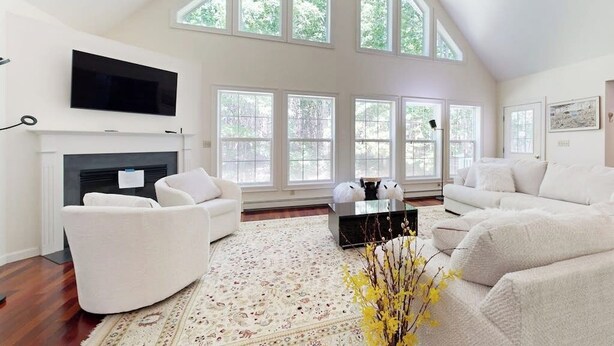 A spacious living room with a fireplace, white sofas, a flat-screen TV, and large windows. - Peaceful Home With Forest Views & Great Yard - Minutes to Town (Wiscasset)
