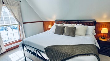 Un dormitorio con cama, almohadas, lámpara de noche, ventana con cortinas y vista al exterior.