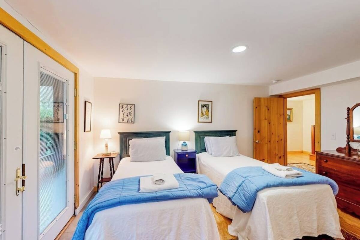 A hotel room with two beds, a wooden wardrobe, a small bedside table with a lamp, and a door with a glass panel.