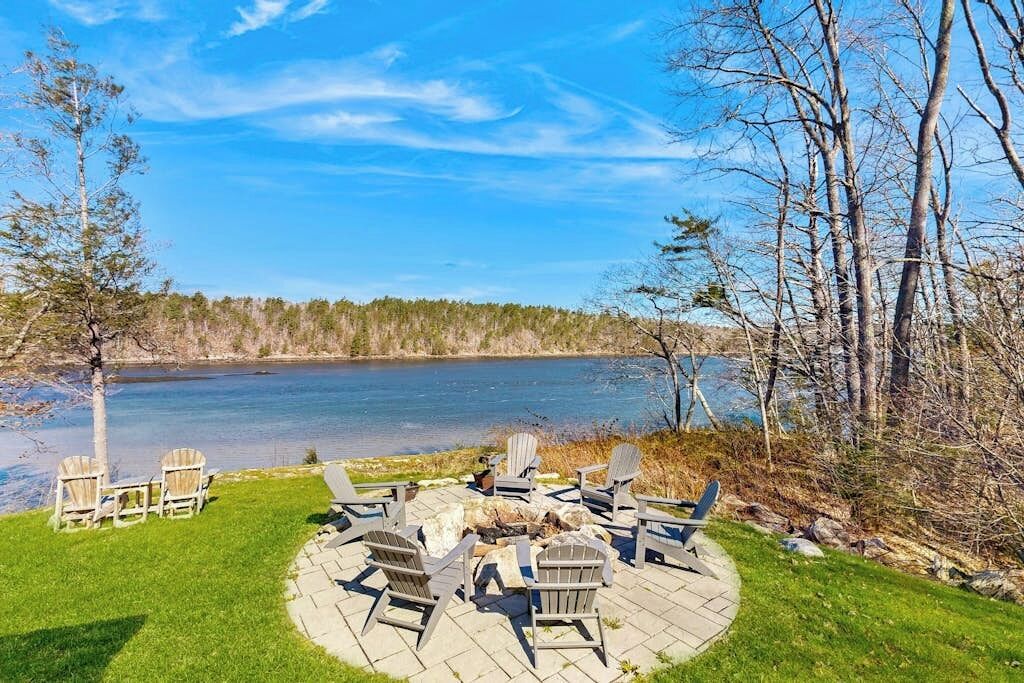 A circular outdoor seating area with Adirondack chairs around a fire pit, overlooking a lake with trees in the background.