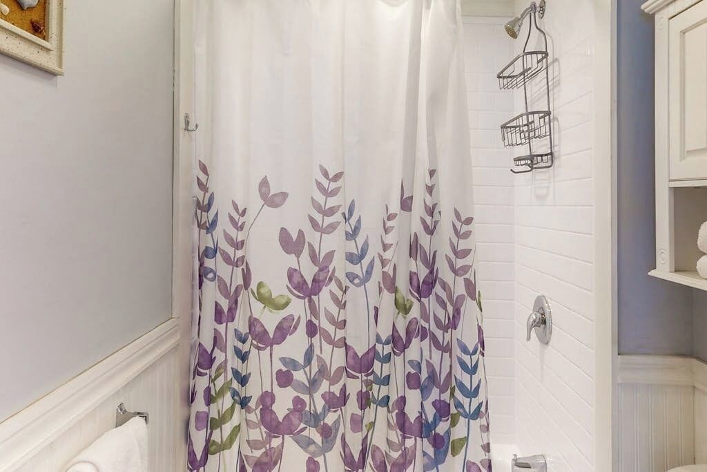 A bathroom with a shower curtain featuring a leaf pattern, a white tiled wall, a towel rack, and a shower caddy.
