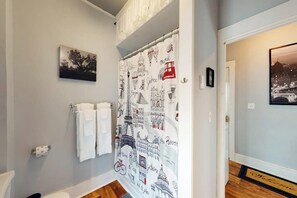 A bathroom with a shower curtain featuring cityscape illustrations, a towel rack with two towels, a framed picture on the wall, and a welcome mat at the entrance. - Corbel House (Bucksport)