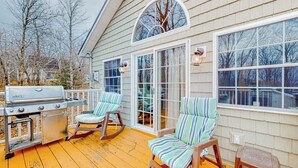 A covered deck with a grill, rocking chairs, and large windows. - Lake-view Two-story Home With Beach Access, Wood Stove, Grill, W/d, & Fast Wifi (Greenville Junction)