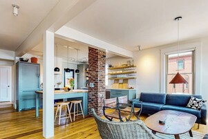 A modern living room with a blue sofa, a wicker chair, a round coffee table, and a kitchen area with a brick column.